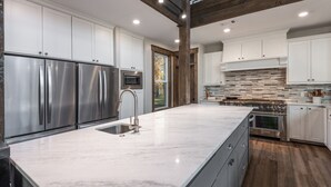 A modern kitchen with stainless steel appliances, white cabinetry, a large island with a sink, and a stone backsplash. - Gorgeous Mountain Lodge on Privately-Owned Lake - Sleeps 30! (Wilburton)