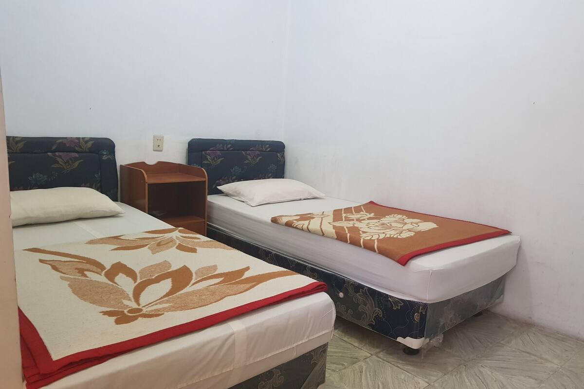 Une chambre avec deux lits, chacun recouvert d’une courtepointe à motifs floraux différents. Une petite table de chevet en bois est placée entre les deux lits.