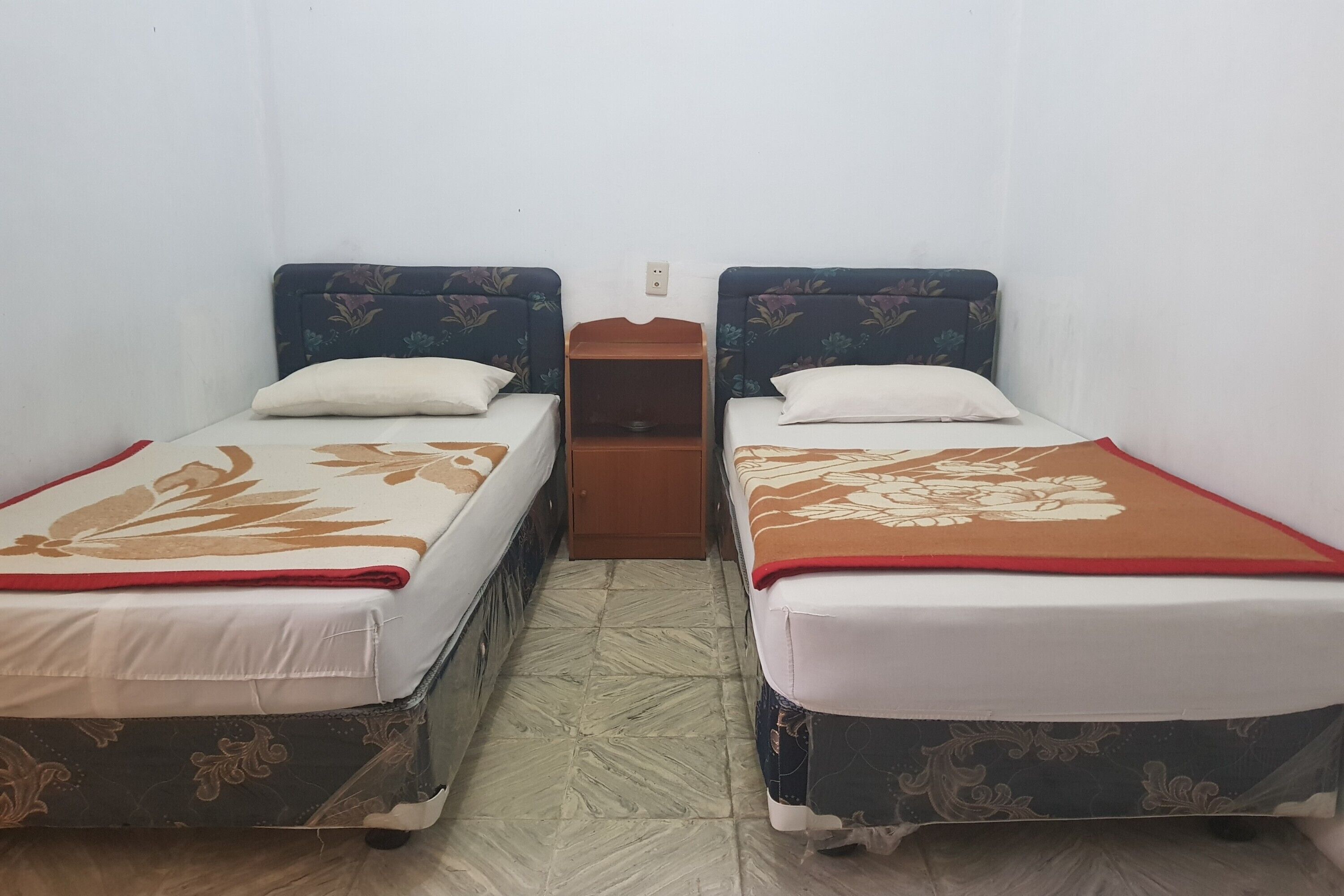 Two single beds with floral-patterned headboards and footboards, a wooden nightstand between them, and a tiled floor.