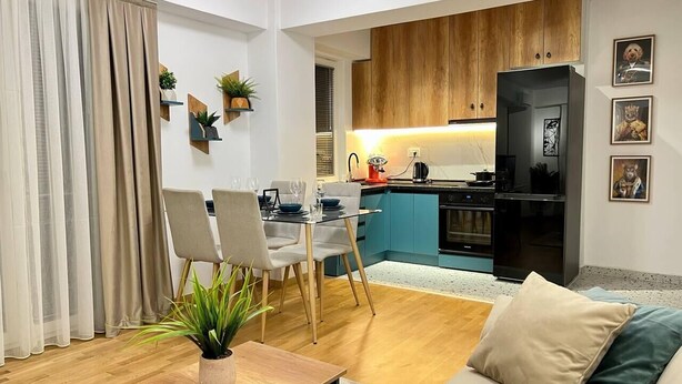 A modern living room with a wooden coffee table, a beige sofa, and a dining area with chairs and a glass table. A kitchen with blue cabinets and a black refrigerator is visible in the background. - Corner penthouse apartment in the centar of Skopje (Skopje)