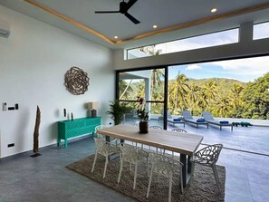 Un espace de repas moderne avec une table et des chaises en bois, une console de couleur turquoise et une vue sur un paysage luxuriant grâce à de grandes fenêtres.
