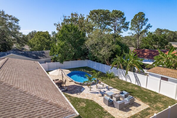 Un patio trasero con piscina, muebles de patio y un área cercada con árboles.