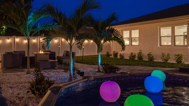 Zona de piscina amb pilotes de colors que floten, un espai d'estar amb mobles d'exterior i un edifici amb finestres i façana ben il·luminada.