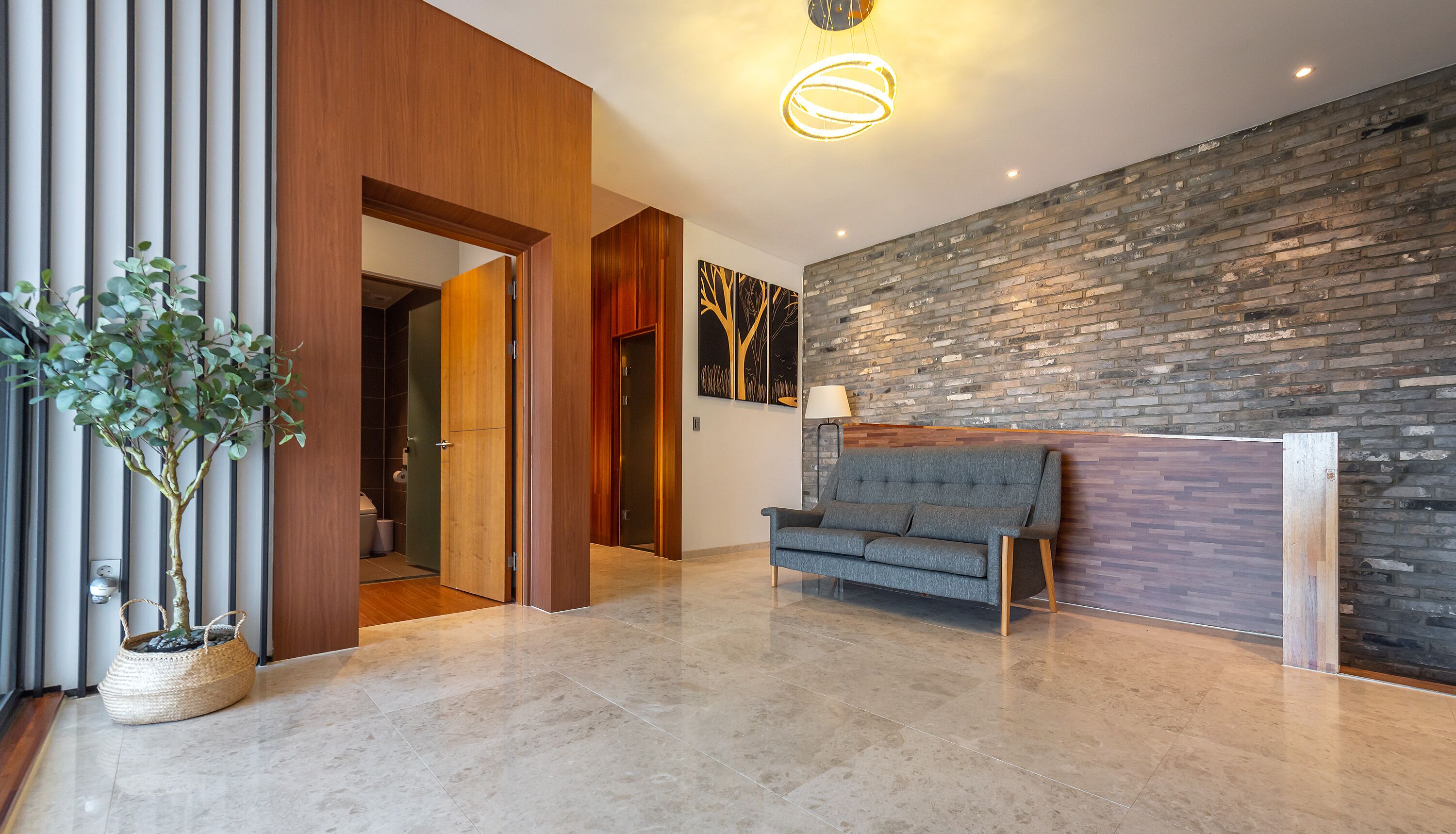 A modern lobby with a grey sofa, a wooden reception desk, a potted plant, and a brick wall.