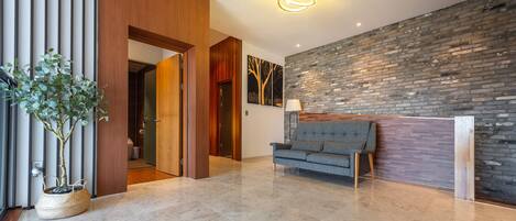 A modern lobby with a grey sofa, a wooden reception desk, a potted plant, and a brick wall.