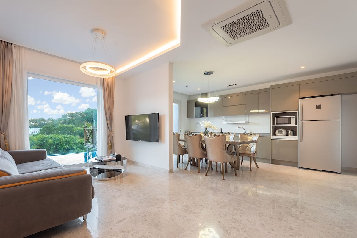 A modern living room with a sofa, dining area, kitchen, and a large window with a view of greenery.