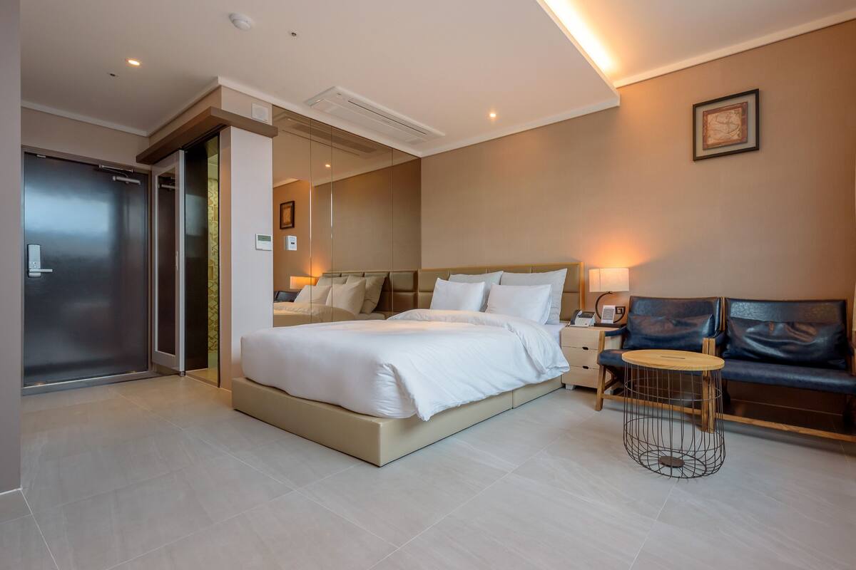 A modern hotel room with a large bed, a small round table, a sofa, and a door leading to a bathroom.