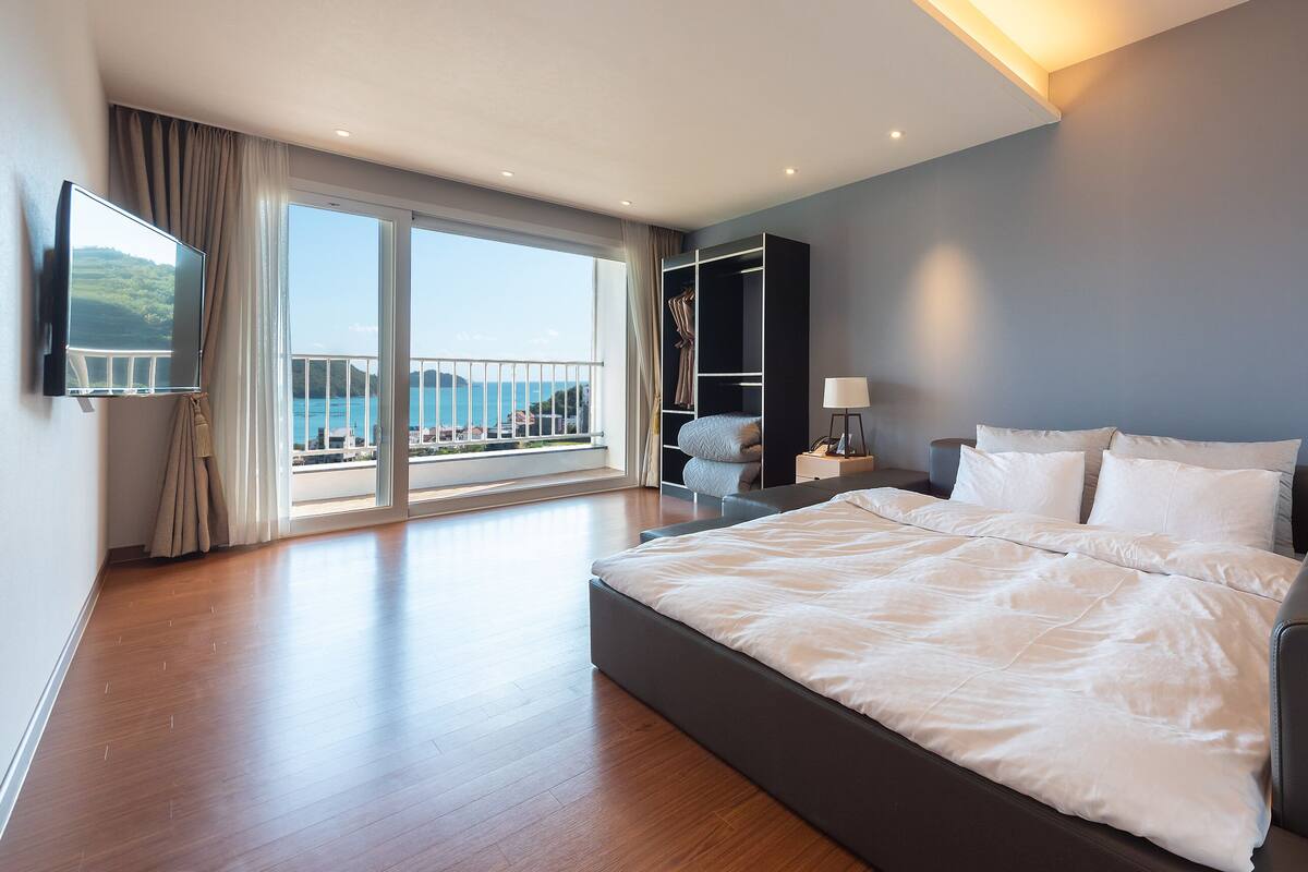 A modern hotel room with a large bed, a bedside table with a lamp, a TV mounted on the wall, and a view of the sea through a balcony.