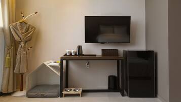 A compact hotel room with a bed, a TV mounted on the wall, a small desk with a coffee maker, a trash bin, and a pet bed.
