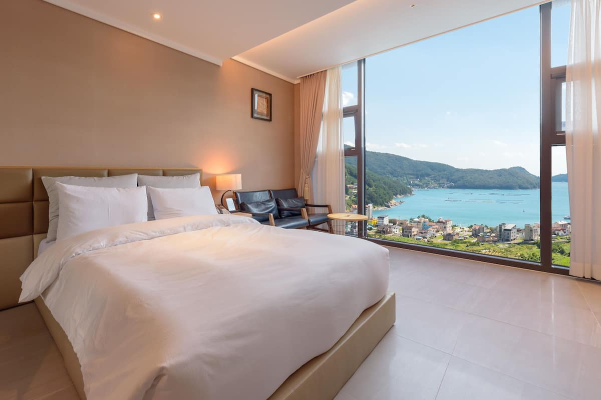 A modern hotel room with a large bed, a chair, a small table, and a view of a coastal town.