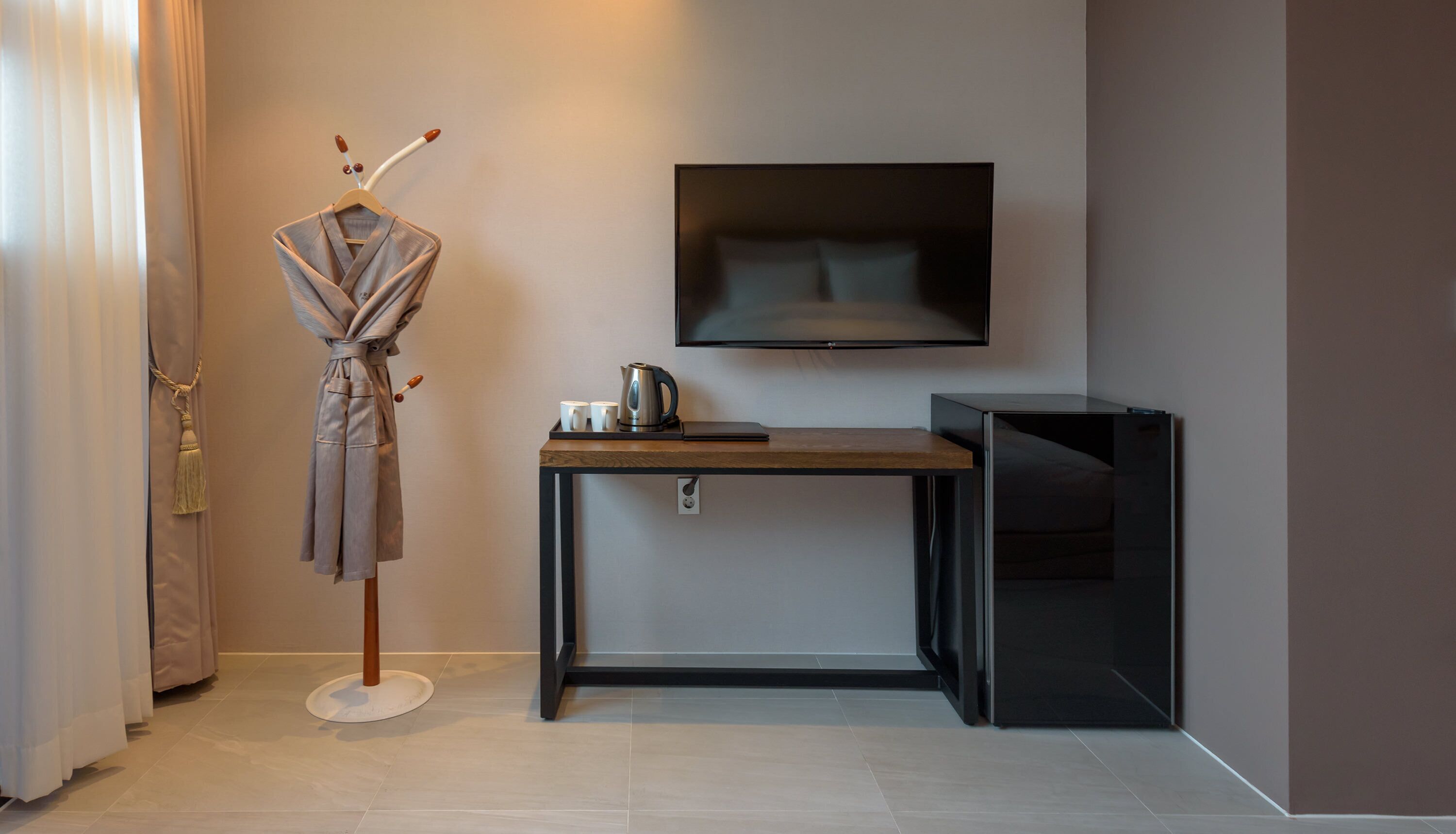 A hotel room with a wooden desk, a flat-screen TV mounted on the wall, a robe hanging on a stand, and a coffee maker on the desk.