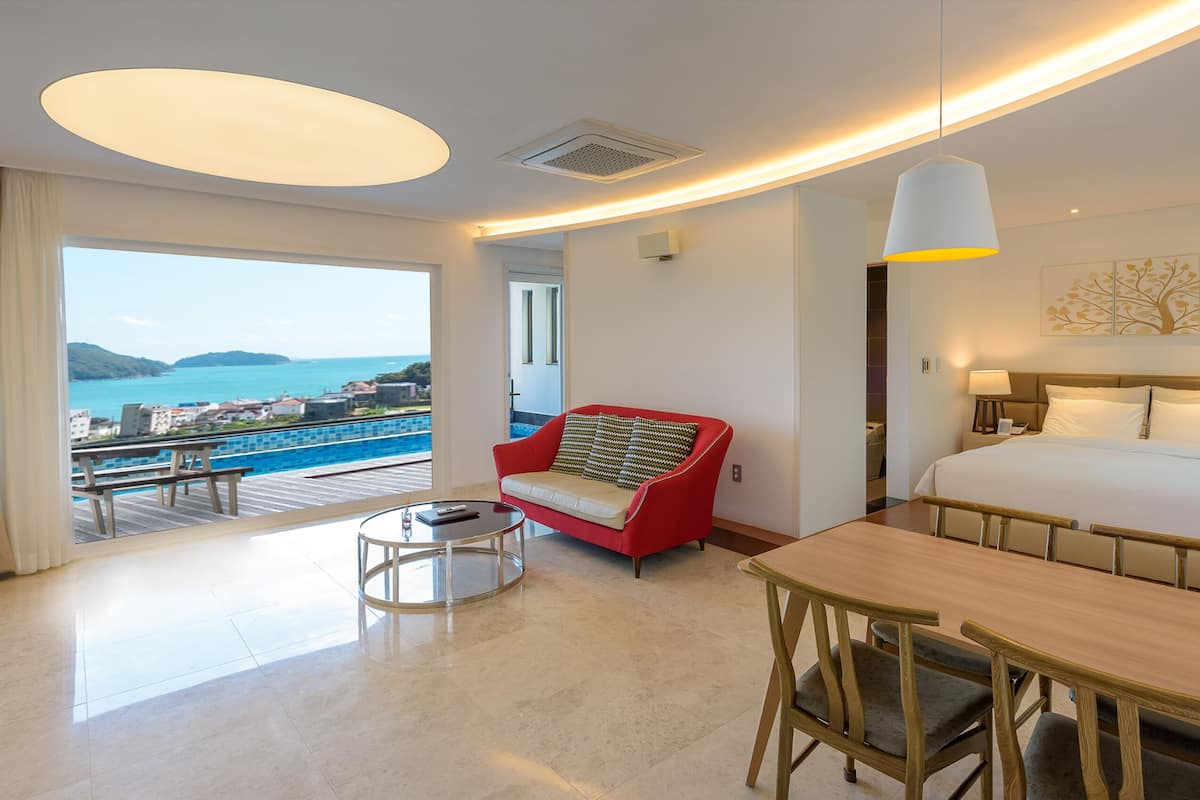 A hotel room with a bed, a red armchair, a round table, a small coffee table, and a view of the sea.
