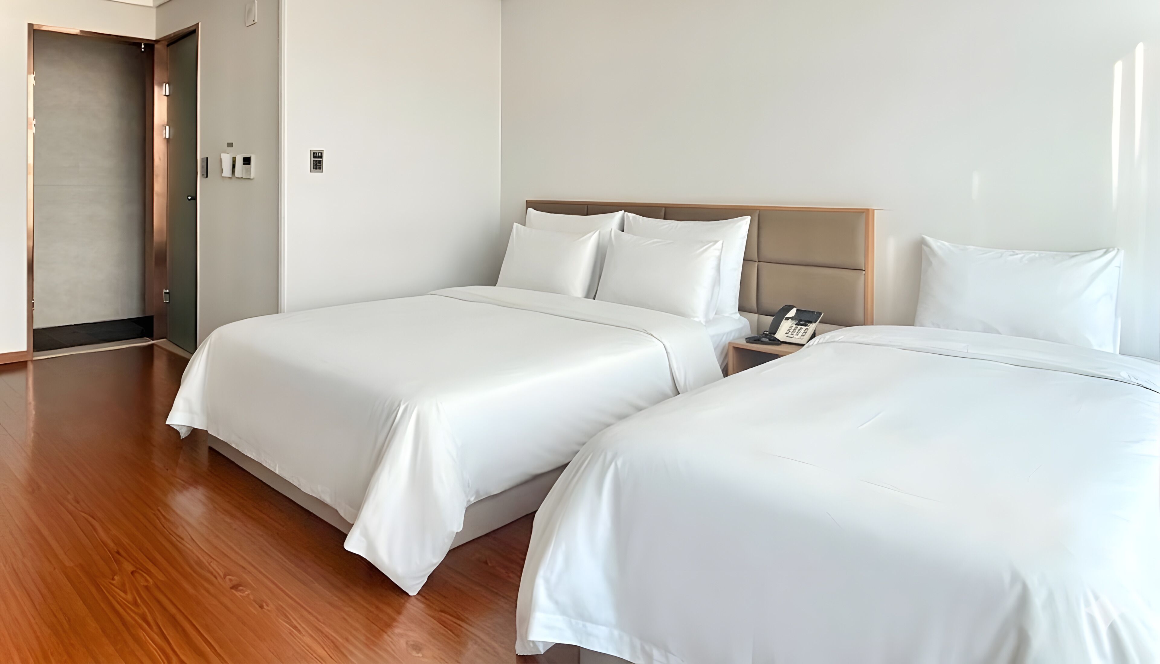 A hotel room with two beds, a wooden floor, a bedside table with a phone, and a door with a light switch.