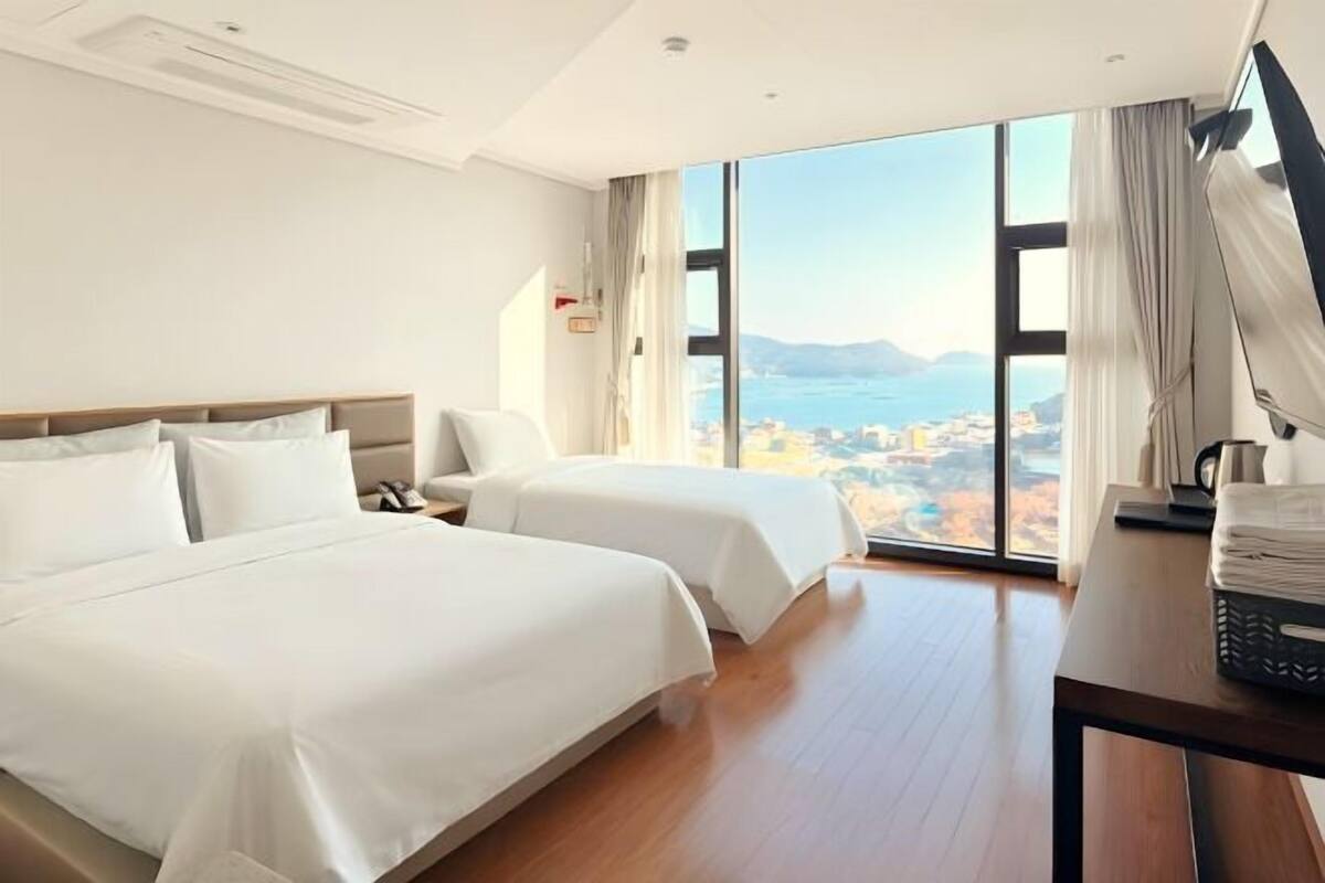 A hotel room with two beds, a desk, a chair, and a large window offering a city view.