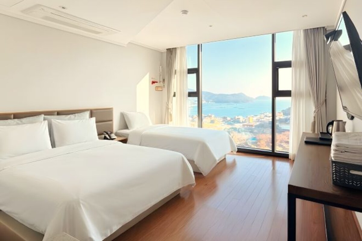 A hotel room with two beds, a desk, a chair, and a large window offering a city view.