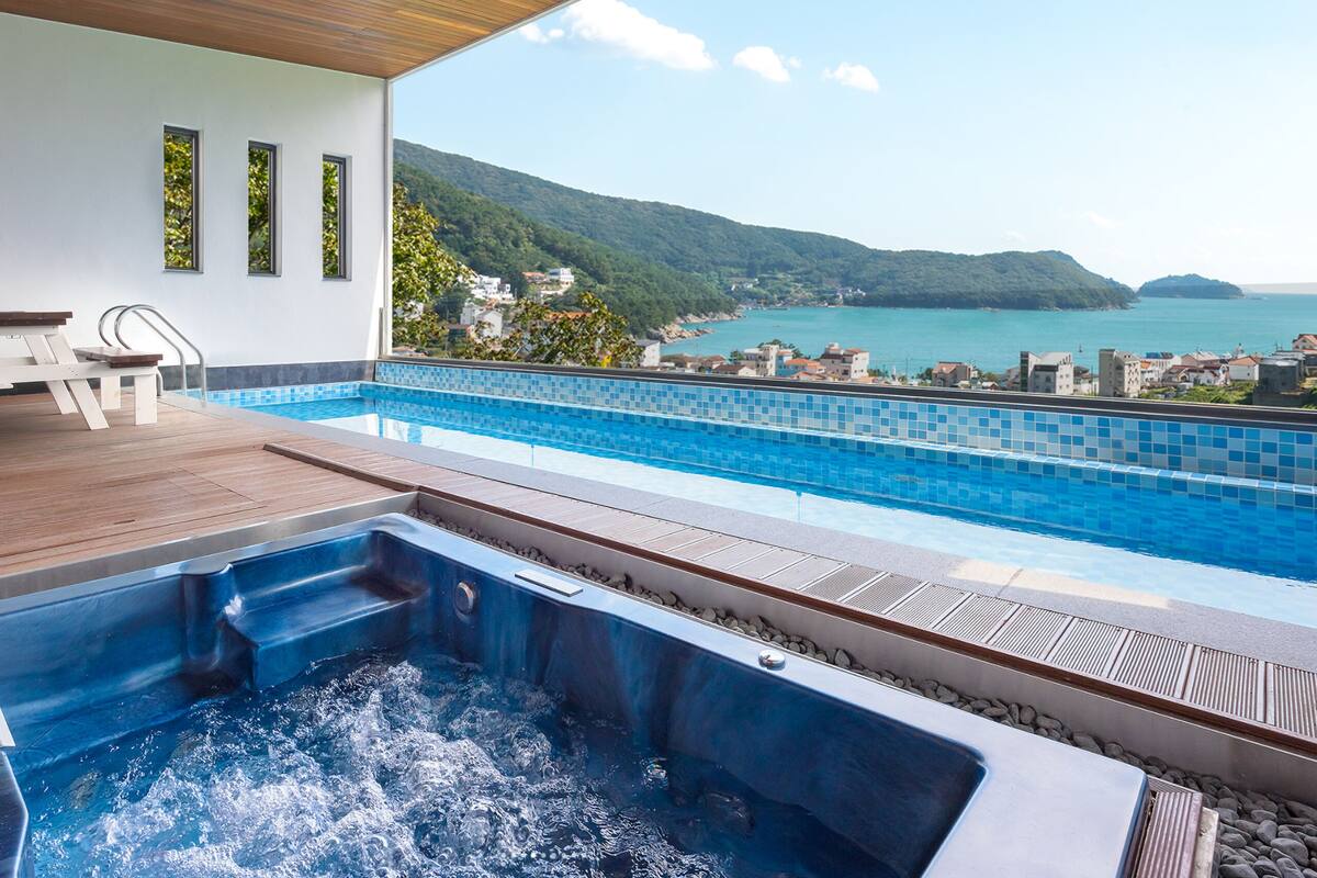A rooftop infinity pool with a hot tub, wooden deck, and a view of the sea and hills.