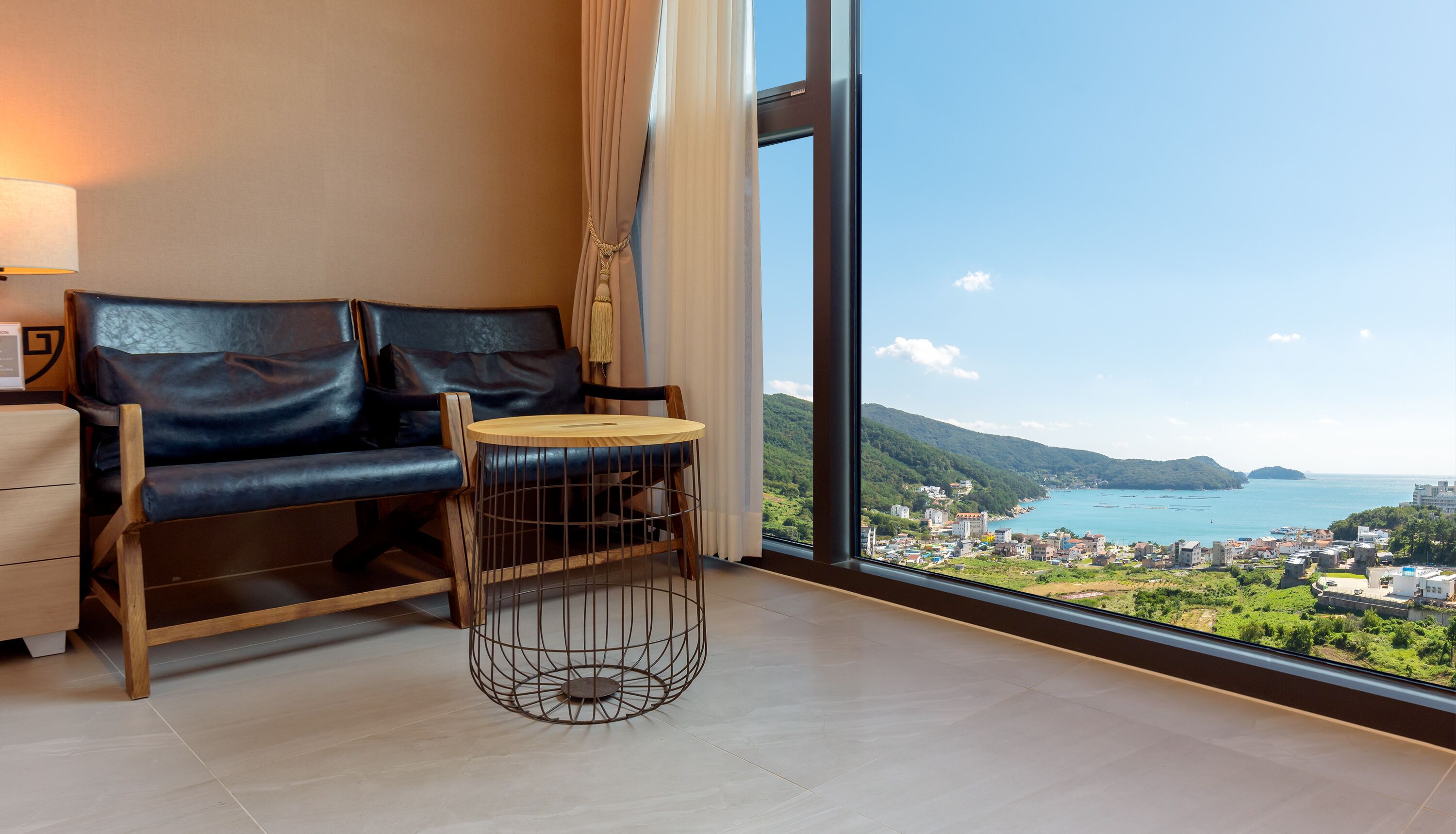Ein modernes Zimmer mit einer Ledersofa, einem kleinen Holztisch und einem großen Fenster mit schöner Aussicht auf eine Küstenstadt und Berge.