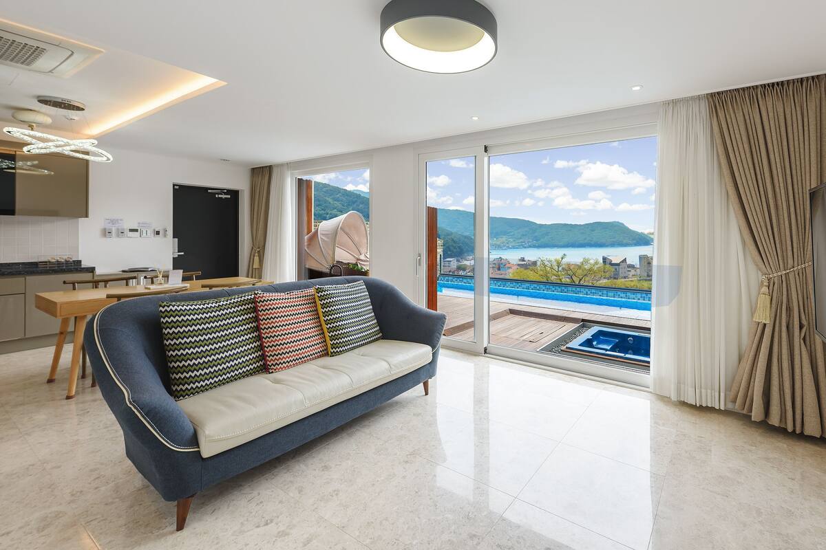 A modern hotel room with a sofa, a dining table, a TV, and a view of a pool and mountains.