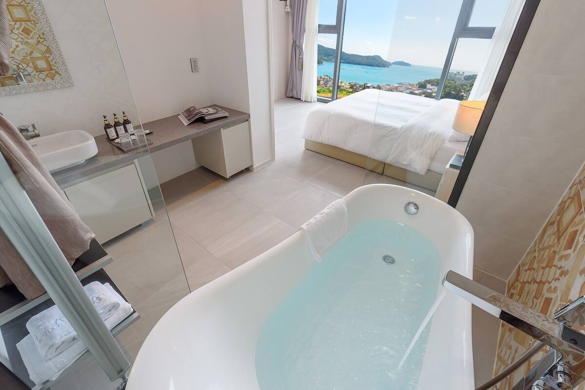 A modern bathroom with a bathtub, a sink, and a mirror. There is a window with a view of the sea and a cityscape.