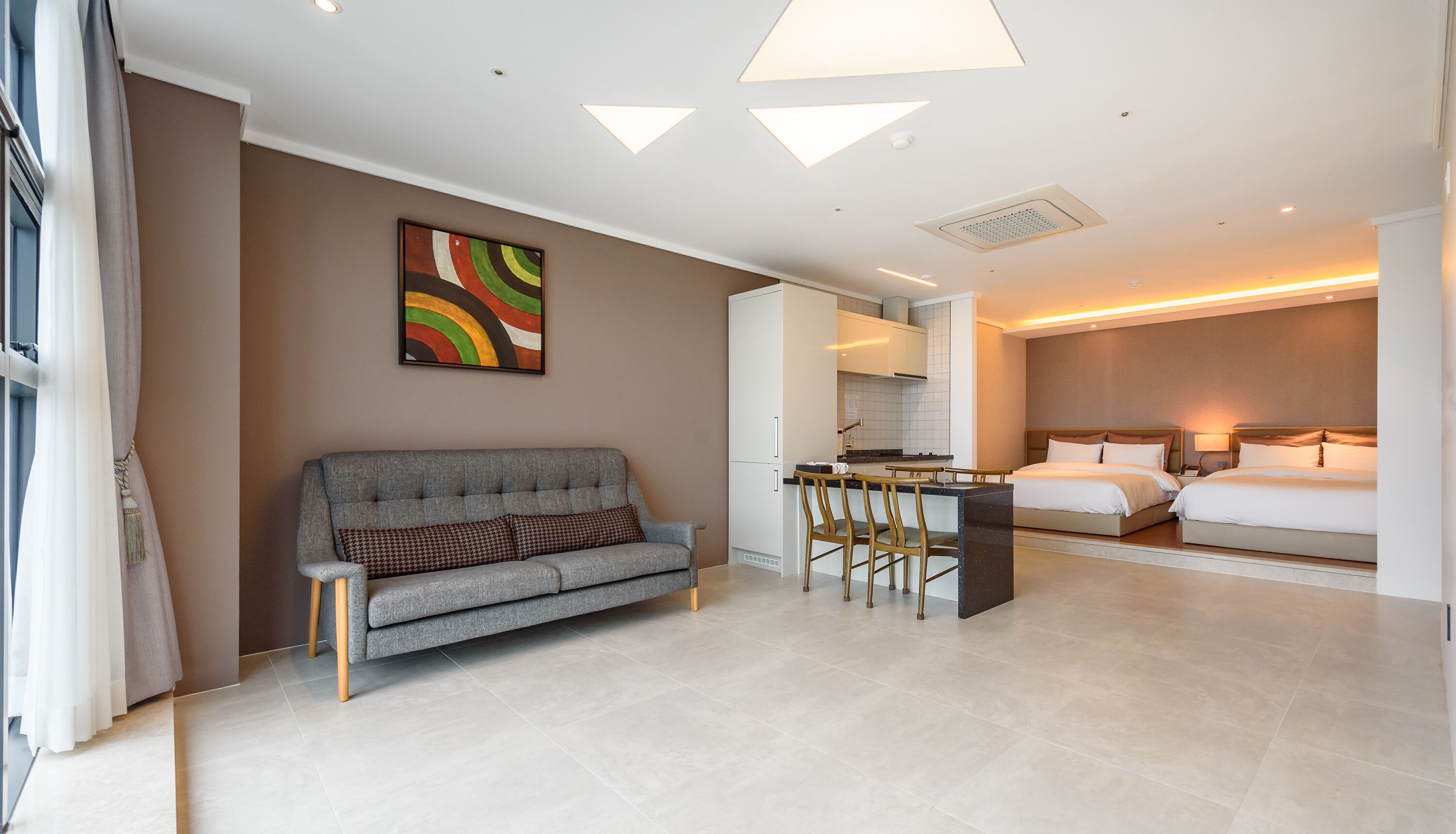 Una habitación de hotel moderna con un sofá gris, una zona de comedor con taburetes de bar, una cama con cabecera de madera y una cocina.