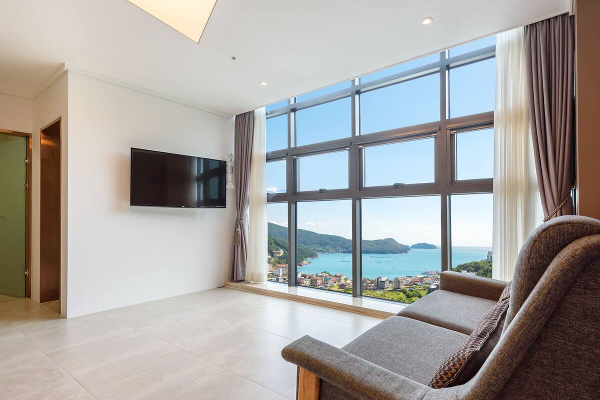 A spacious room with a large window offering a view of a coastal area, a flat-screen TV mounted on the wall, and a comfortable seating area with a sofa and armchair.