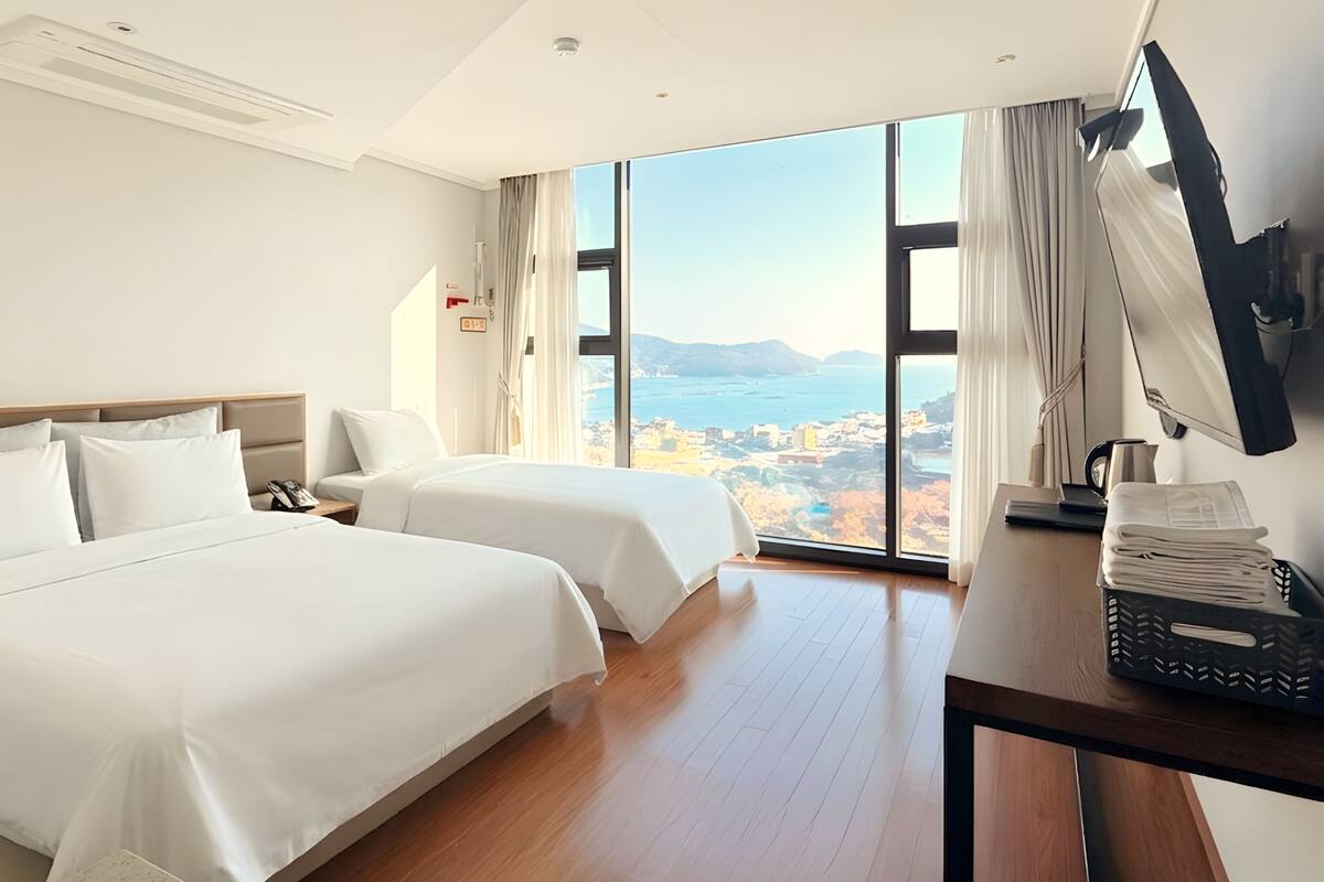 A hotel room with two beds, a desk with a computer, a TV, and a large window with a view of the sea and hills.