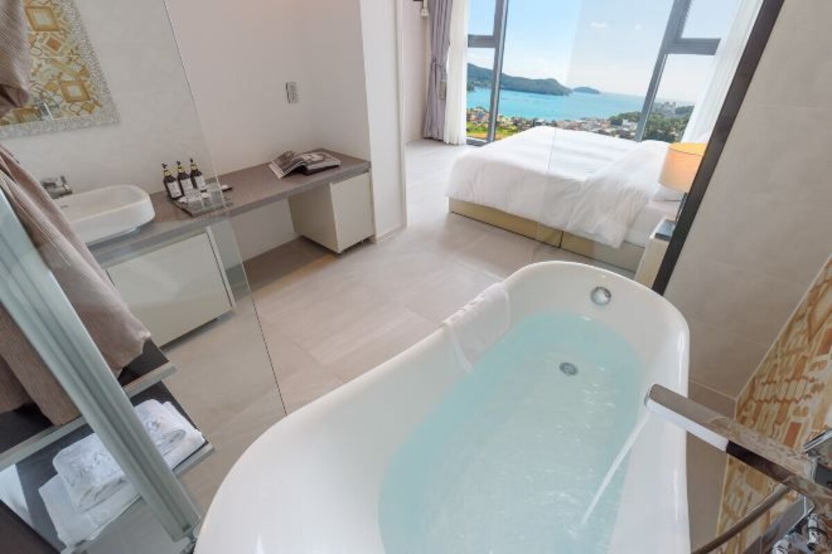 A modern bathroom with a bathtub, a large window, and a view of the sea.