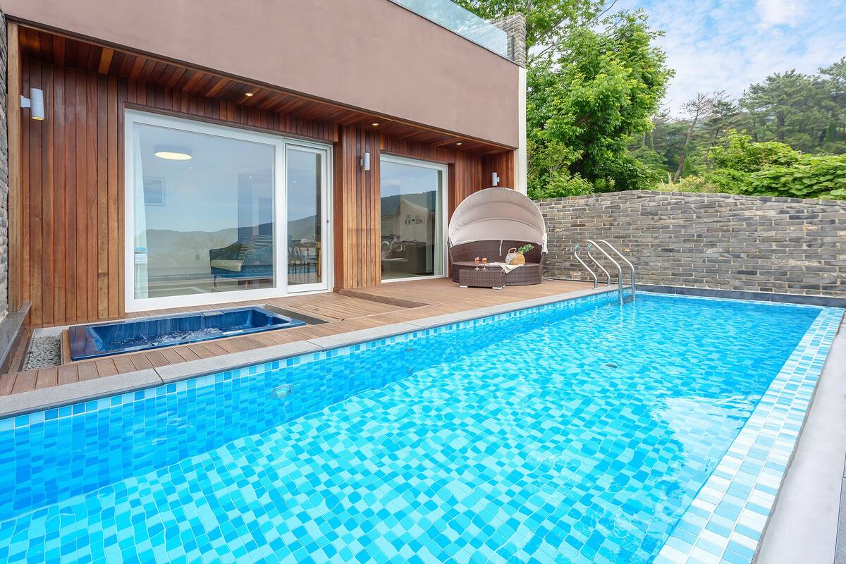 A modern outdoor pool area with a wooden deck, a built-in hot tub, and a stone wall.