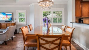 A dining area with a large table, six chairs, a chandelier, and a television on a stand.