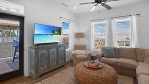 A living room with a television displaying a beach scene, a beige sofa, a round woven ottoman, and a wooden cabinet. - Shellfish Cottage offers gulf views, pool, hot tub, outdoor kitchen! Steps to beach! (Port Saint Joe)