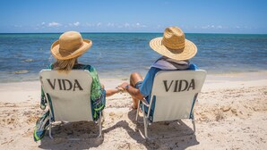 Two people sitting on beach chairs facing the ocean, with the word "VIDA" on the backrests. - VIDA Cayman (West Bay)