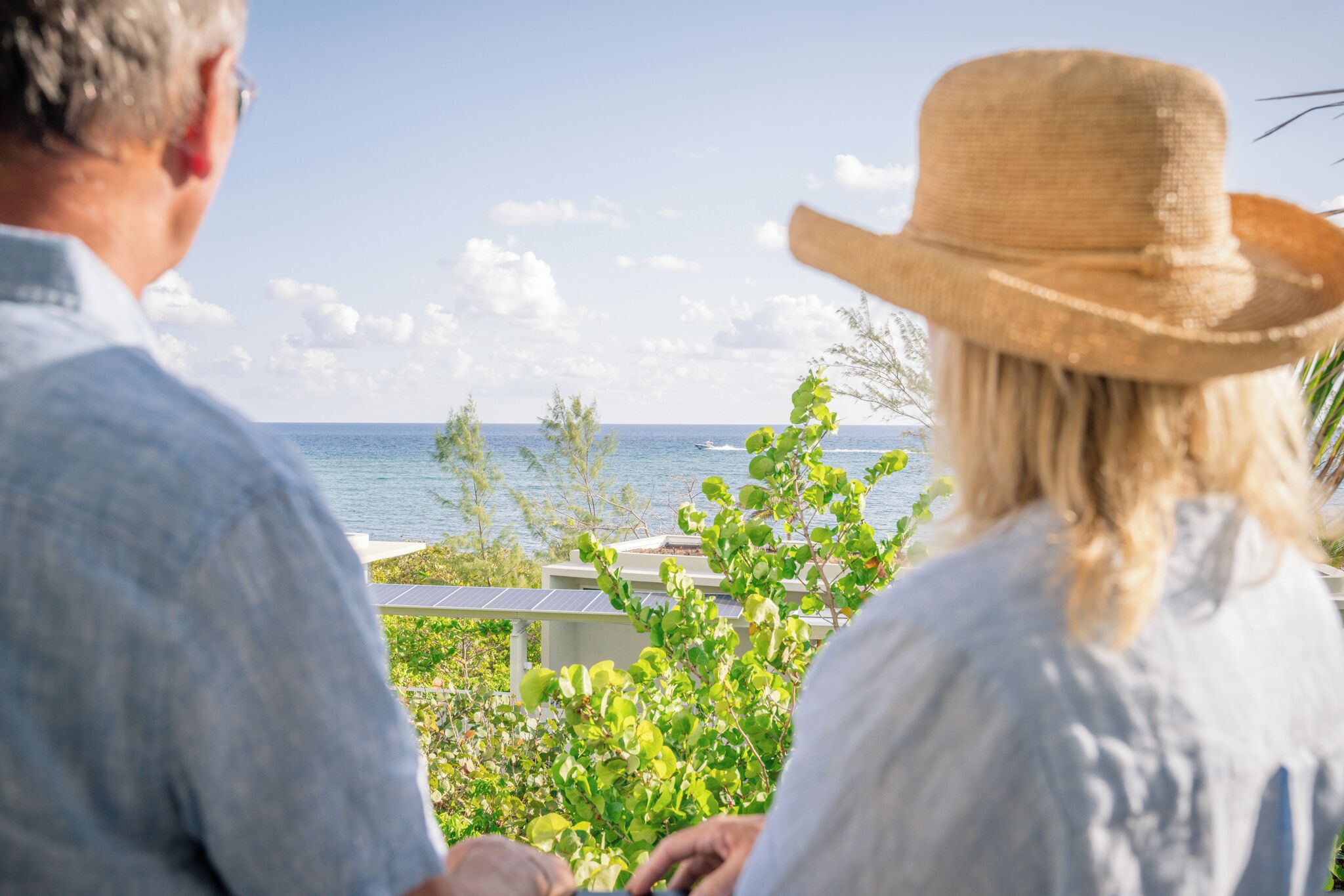 A couple seated on a balcony overlooking a body of water, with greenery and a clear sky.