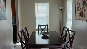 Un comedor con una mesa rectangular, seis sillas, un bol en la mesa, una lámpara en el techo y una ventana con persianas.