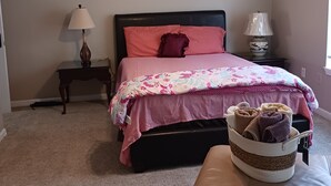 Un dormitorio con una cama, un ventilador de techo, dos mesitas de noche, una lámpara y una cesta de toallas.