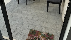 Une terrasse extérieure avec une table ronde et six chaises, un paillasson orné de motifs floraux et, en arrière-plan, une vue sur une maison.