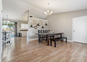 Un comedor amplio con una mesa de madera, sillas y un banco. Hay estantes en la pared con plantas y fotografías enmarcadas. Un candelabro cuelga del techo, y se ve una puerta blanca a la derecha.