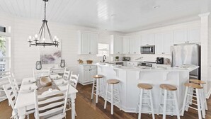 A modern kitchen with white cabinetry, a central island with stools, a chandelier, and a dining area with white chairs. - Better Together by Brunswick Vacation Rentals: Heated Pool, Great Views & Access (Ocean Isle Beach)
