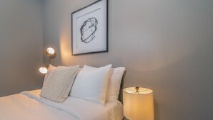 A neatly made bed with white linens and pillows, a wall-mounted abstract art piece, and a bedside lamp with a wooden base.