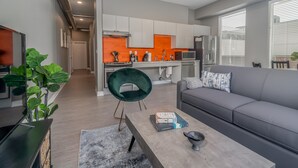 A modern living room with a grey sofa, a coffee table with books, a green chair, and a kitchen in the background.