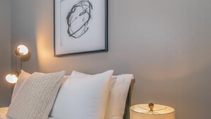Un lit bien fait, avec du linge de lit blanc et des oreillers, une œuvre d’art abstraite encadrée accrochée au mur, et une lampe de table à la base en forme de nid d’abeilles, placée à côté du lit.