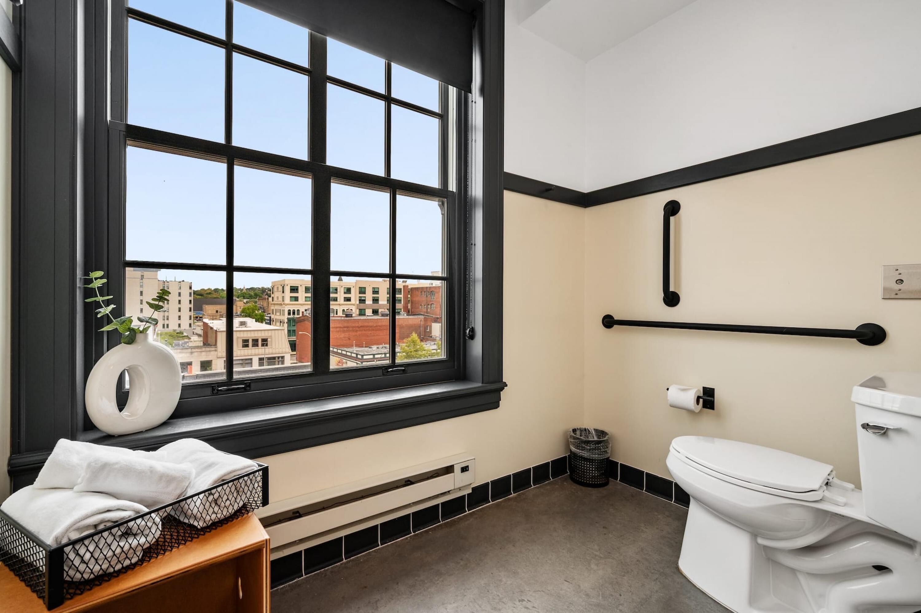 A bathroom with a large window, a toilet, a towel rack, and a trash bin.