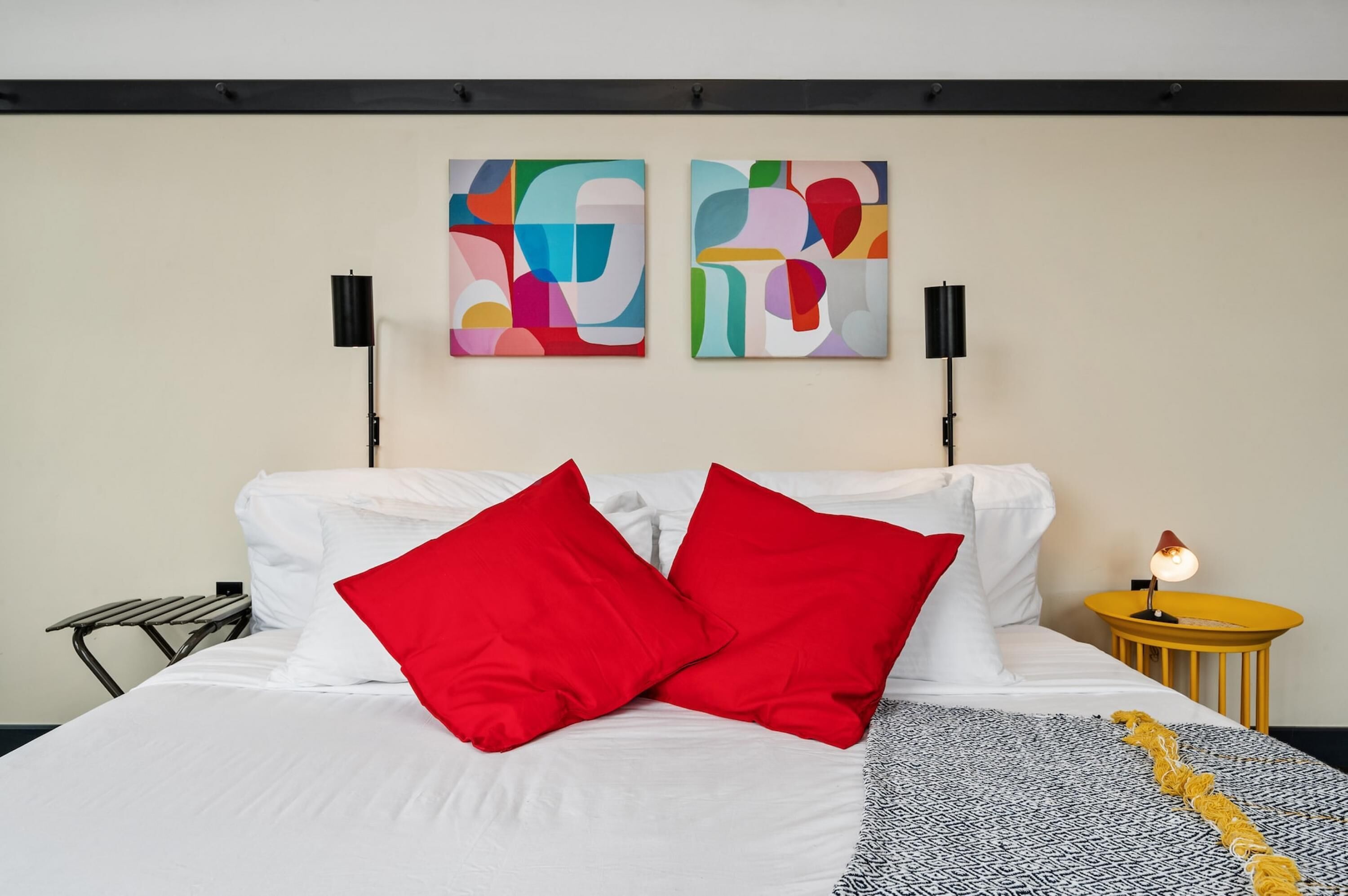 Een net opgemaakt bed met witte en rode kussens, een gebreid plaid en twee abstracte schilderijen aan de muur.