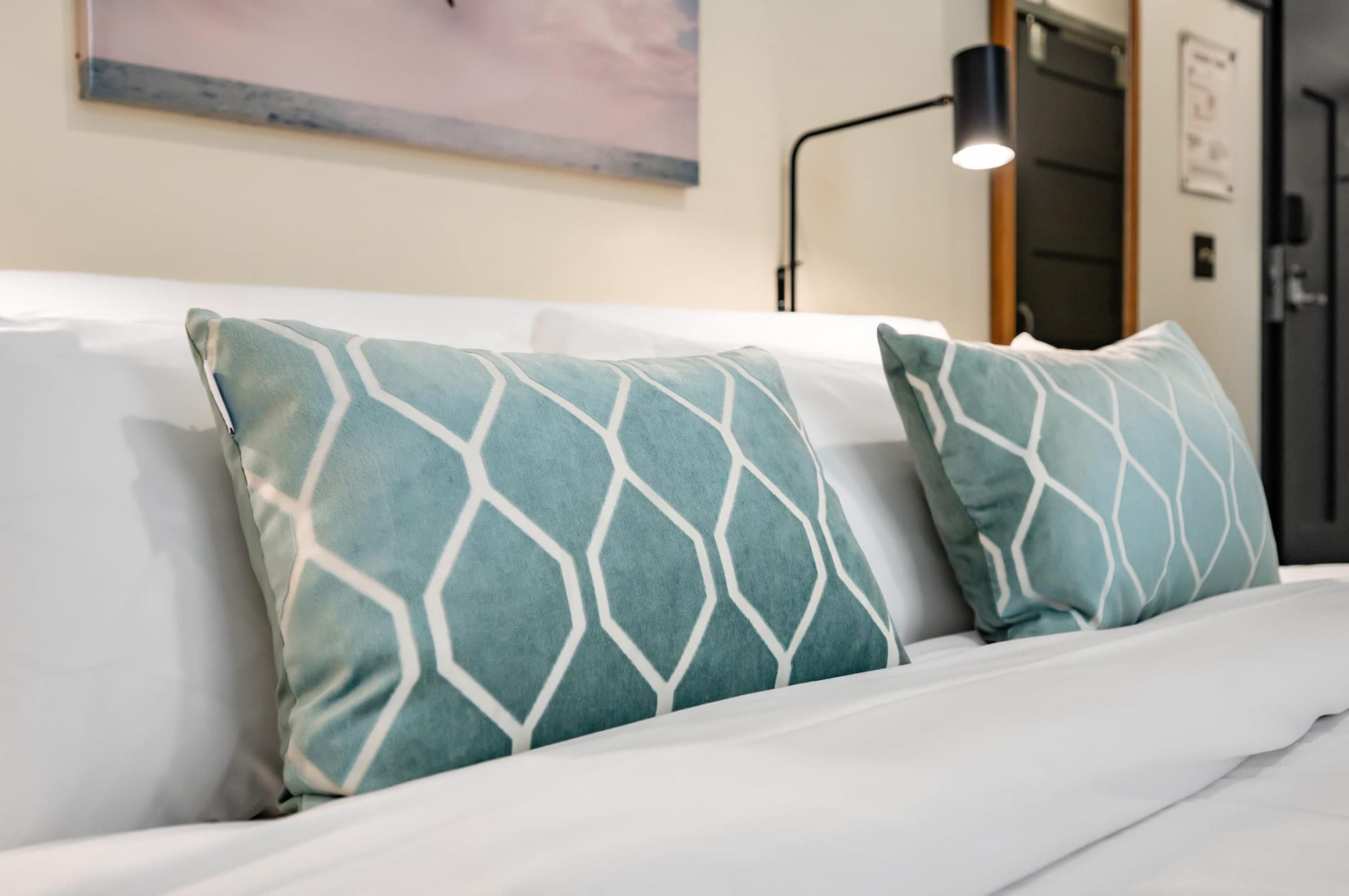 A bed with white bedding and two teal pillows with a geometric pattern.