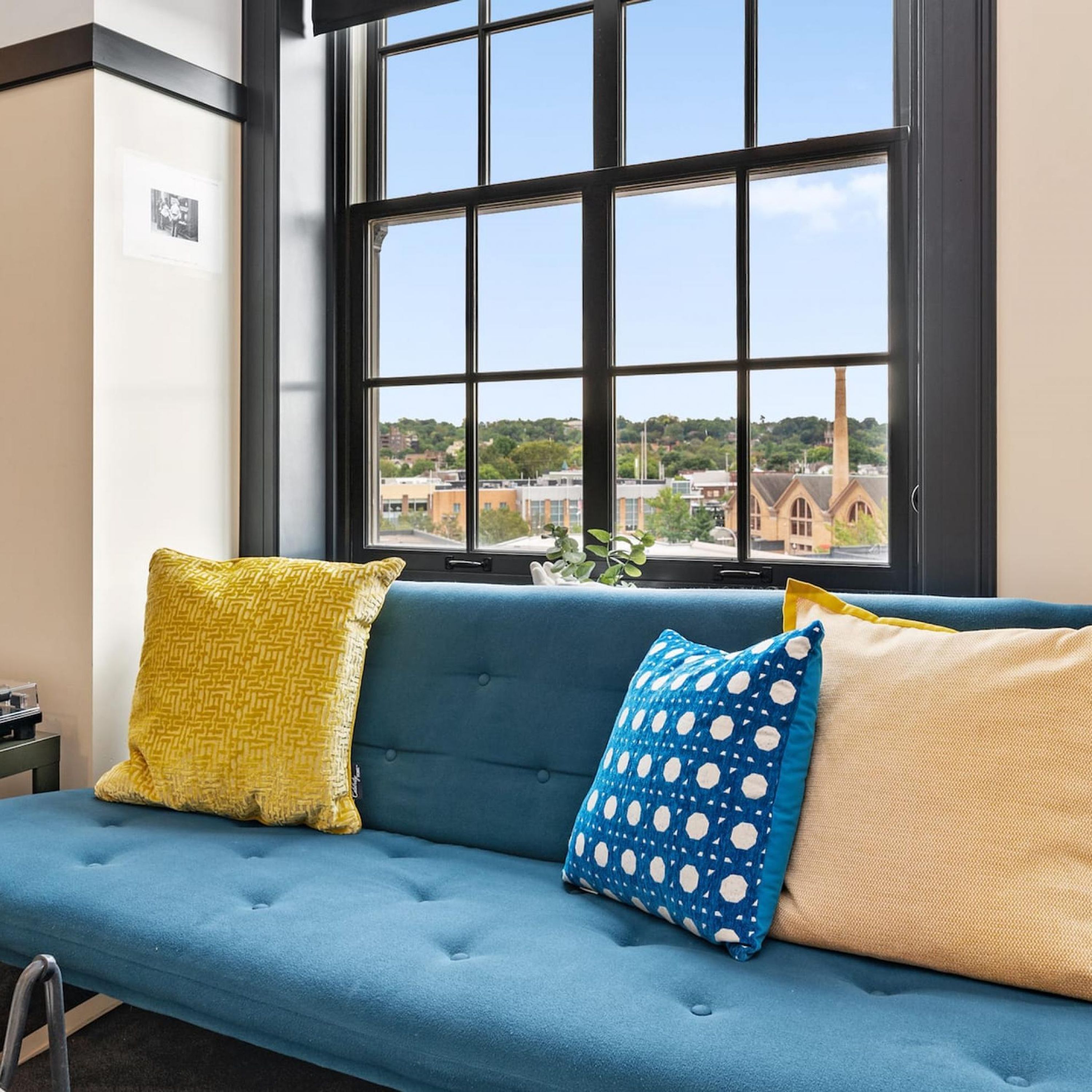 A blue sofa with patterned cushions, a window with a city view, and a small plant on the windowsill.