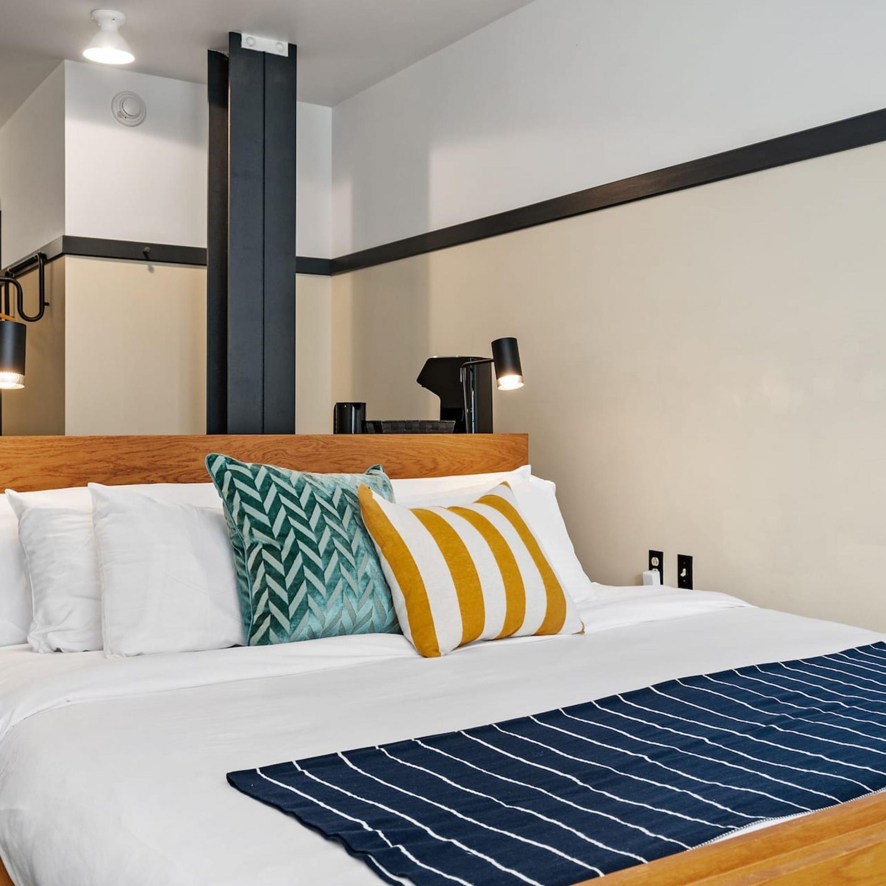 A hotel room with a bed, pillows, a bedside table, a lamp, and a wall-mounted light fixture.