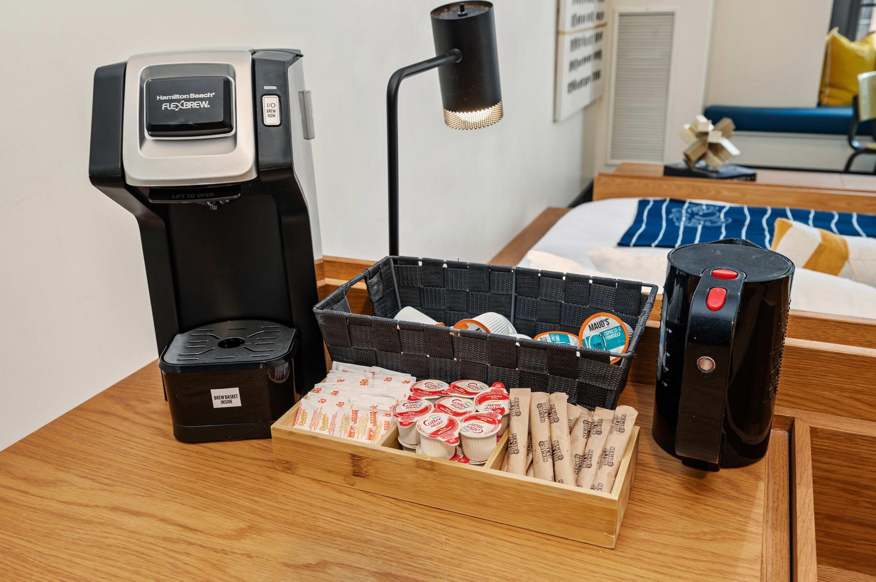 A coffee maker, a lamp, a basket with coffee pods, a coffee machine, and a tray with coffee cups on a wooden table.