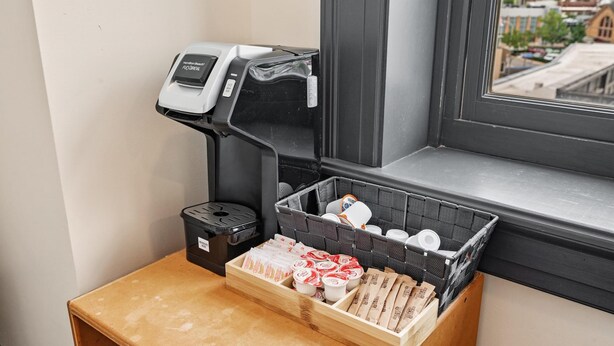 A coffee machine, a trash bin, and a basket with packaged goods on a small table by a window. - Kasa Comfy Studio Nearby Bakery Square The Maverick (Pittsburgh)