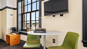 A room with a large window, a white table, two green chairs, a small orange stool, and a television mounted on the wall. - Kasa Comfy Studio Nearby Bakery Square The Maverick (Pittsburgh)