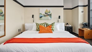 A hotel room with a large bed, white bedding, an orange throw blanket, a green and white patterned pillow, a painting on the wall, and a wooden nightstand with a lamp. - Kasa Comfy Studio Nearby Bakery Square The Maverick (Pittsburgh)