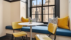A corner seating area with a round table and two yellow chairs, a blue cushioned bench with patterned pillows, and a view of historic architecture through a large window.
